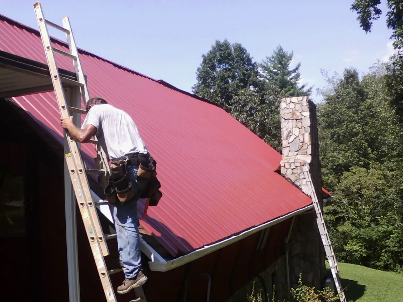 Roofer on a ladder beside a red metal roof and stone chimney — Soffit & Fascia Repair by Roofing Brothers in Wood Dale